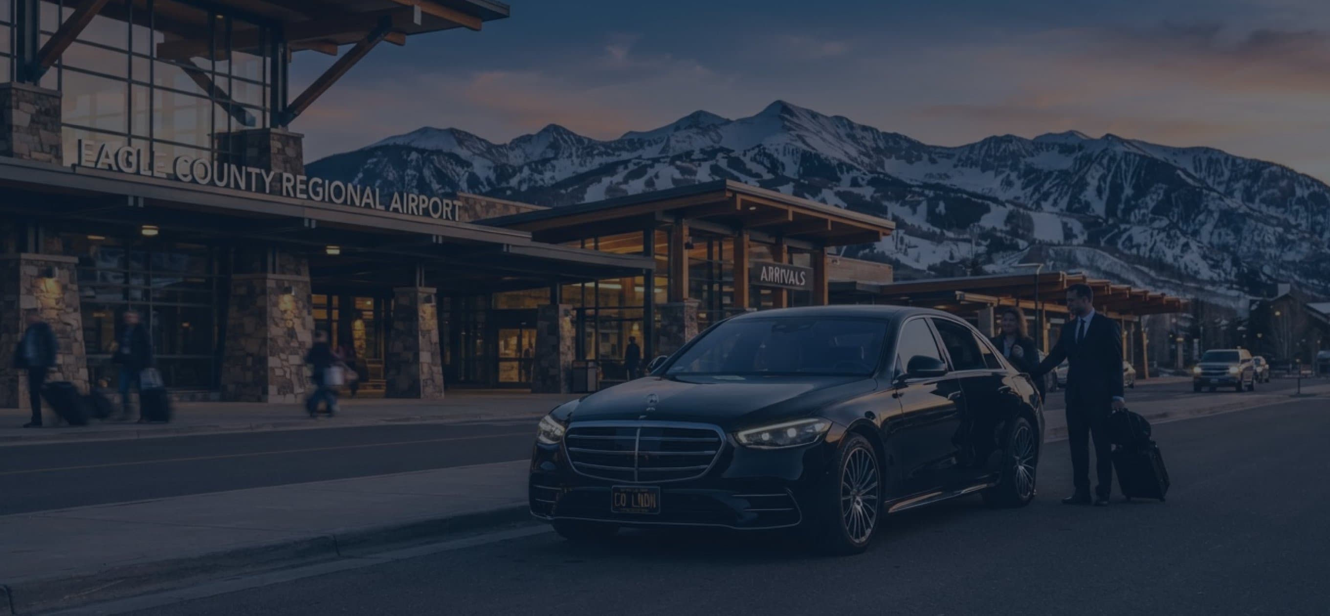 A chauffeur with a black sedan outside a mountain airport terminal at dusk